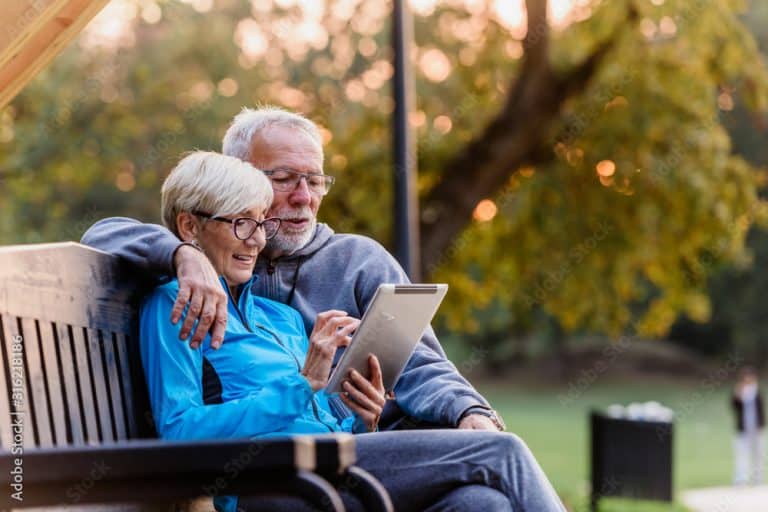 Smiling senior active couple sitting on the bench looking at tablet computer. Using modern technology by elderly.